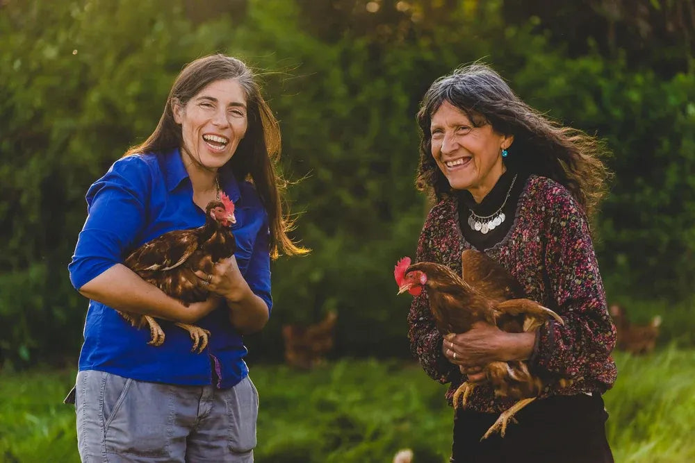Dos mujeres EcoFarmers sonriendo y sosteniendo gallinas libres a pastoreo en la pradera, representando el comercio justo y el bienestar animal en Ecoterra Chile.