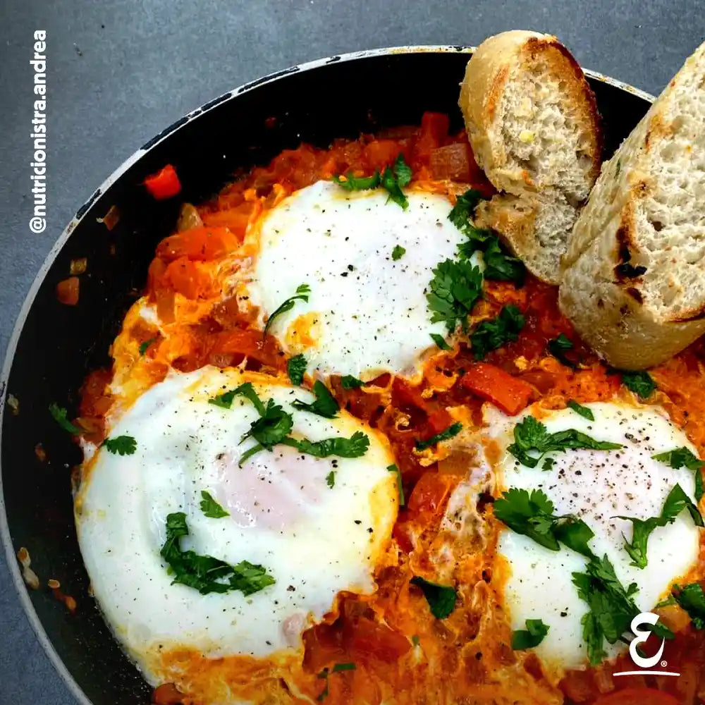 Receta de Shakshuka en sartén con salsa de tomate y tres huevos Ecoterra de gallinas libres de pastoreo, decorada con perejil y tostadas de pan.